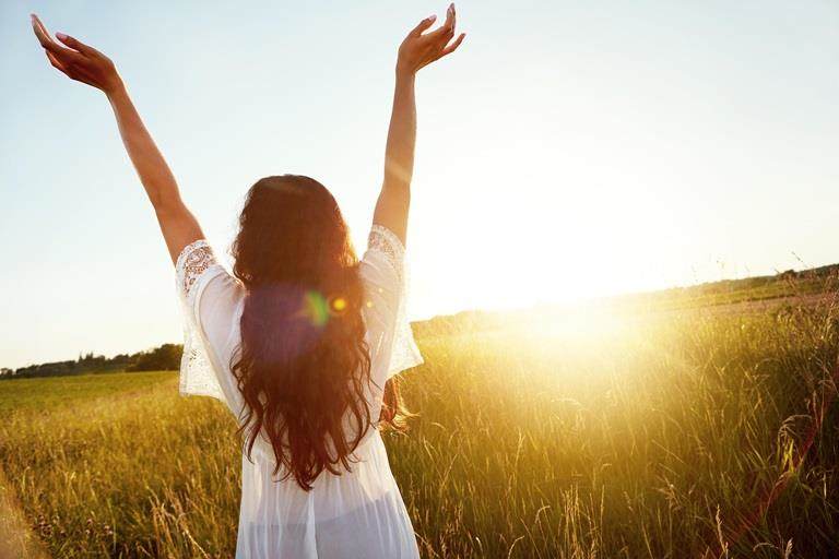 Young woman in field wearing floaty white dress with arms raised